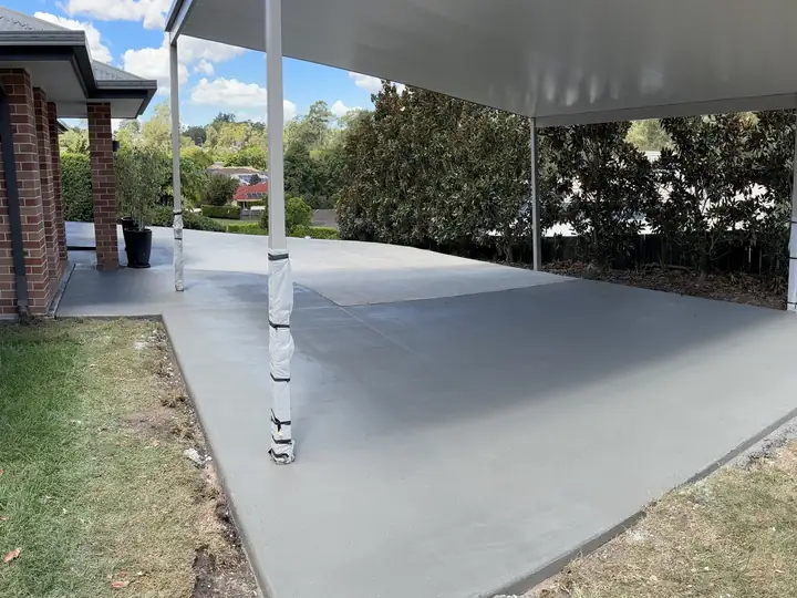 A wide new concrete slab under a carport roof beside a red brick house and lawn