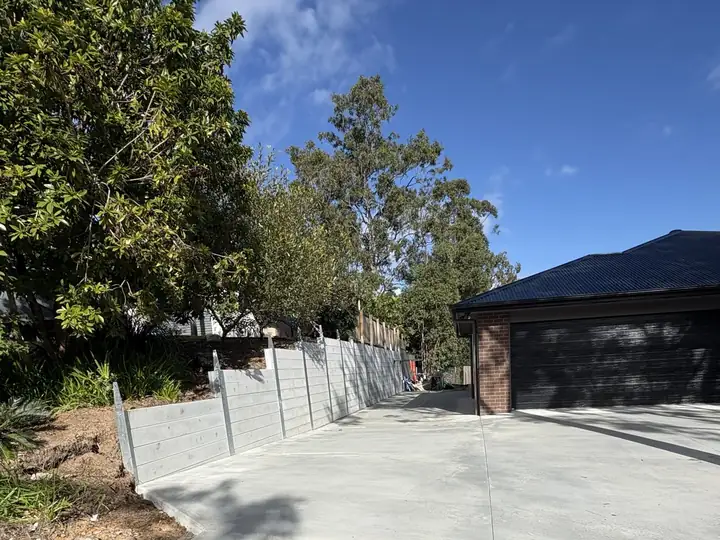 A new plain concrete driveway curving up to a dark brick home in Karalee, with a light concrete sleeper wall along one side