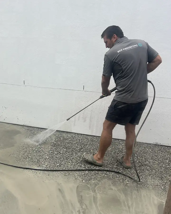 A DCF worker in a grey DCF Concreting shirt pressure-washing fresh exposed aggregate beside a white wall, the stone coming up where the spray lands
